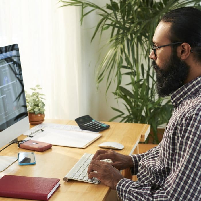 Contemporary programmer in casualwear sitting by desk in front of computer monitor while developing new software